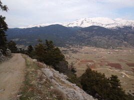 Paisagem montanhosa com um pico nevado ao fundo, um caminho de terra subindo e edifícios espalhados no vale abaixo.