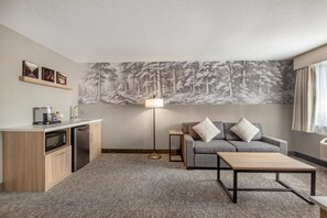 A modern hotel room with a kitchenette, a gray sofa with two pillows, a standing floor lamp, and a large forest mural on the wall.