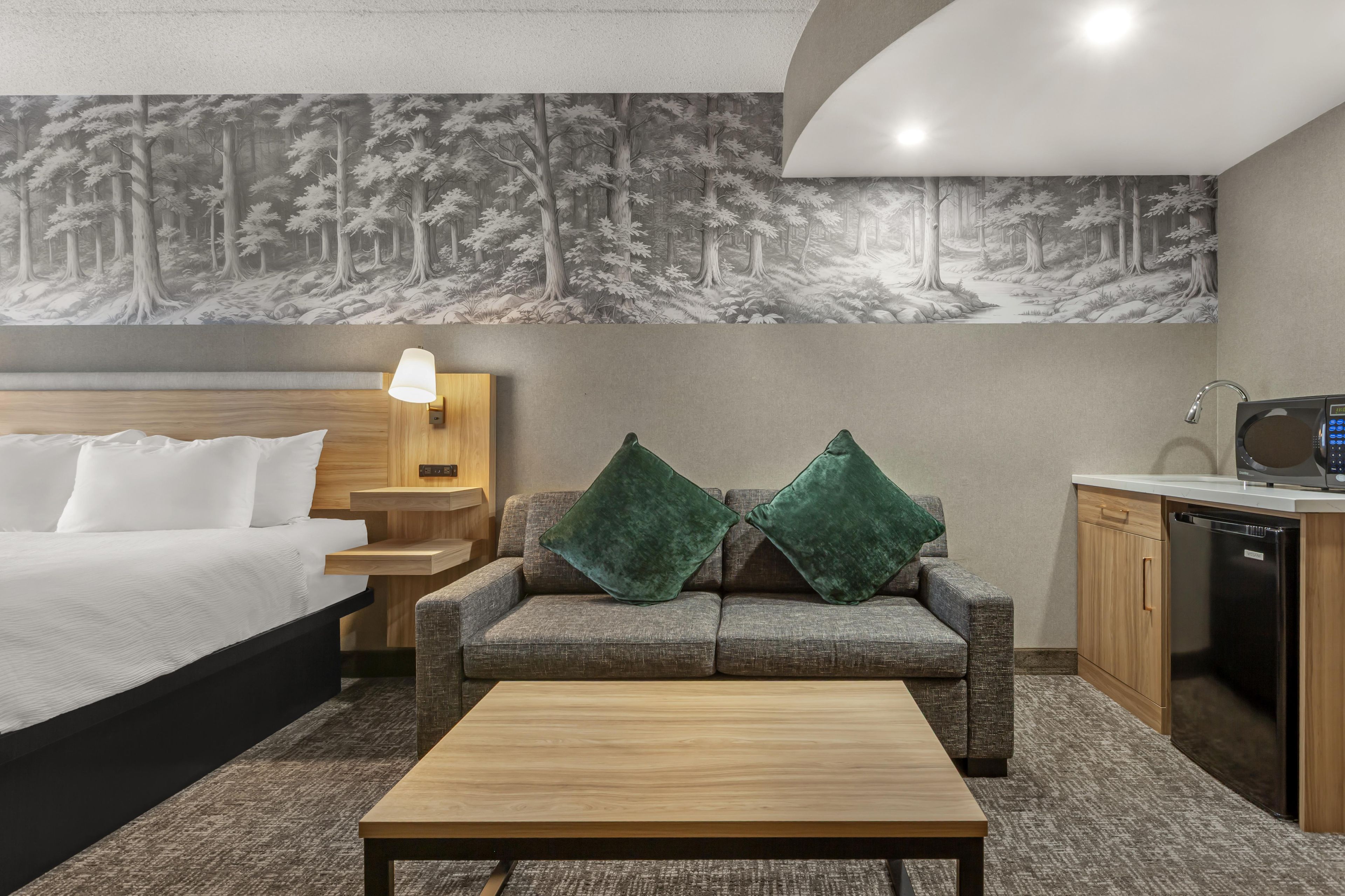 A hotel room with a bed, a sofa with two green cushions, a wooden coffee table, a nightstand with a lamp, and a microwave on a counter.