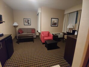 A hotel room with a checkered carpet, a sofa, a red armchair, a coffee table, a TV, and a lamp.