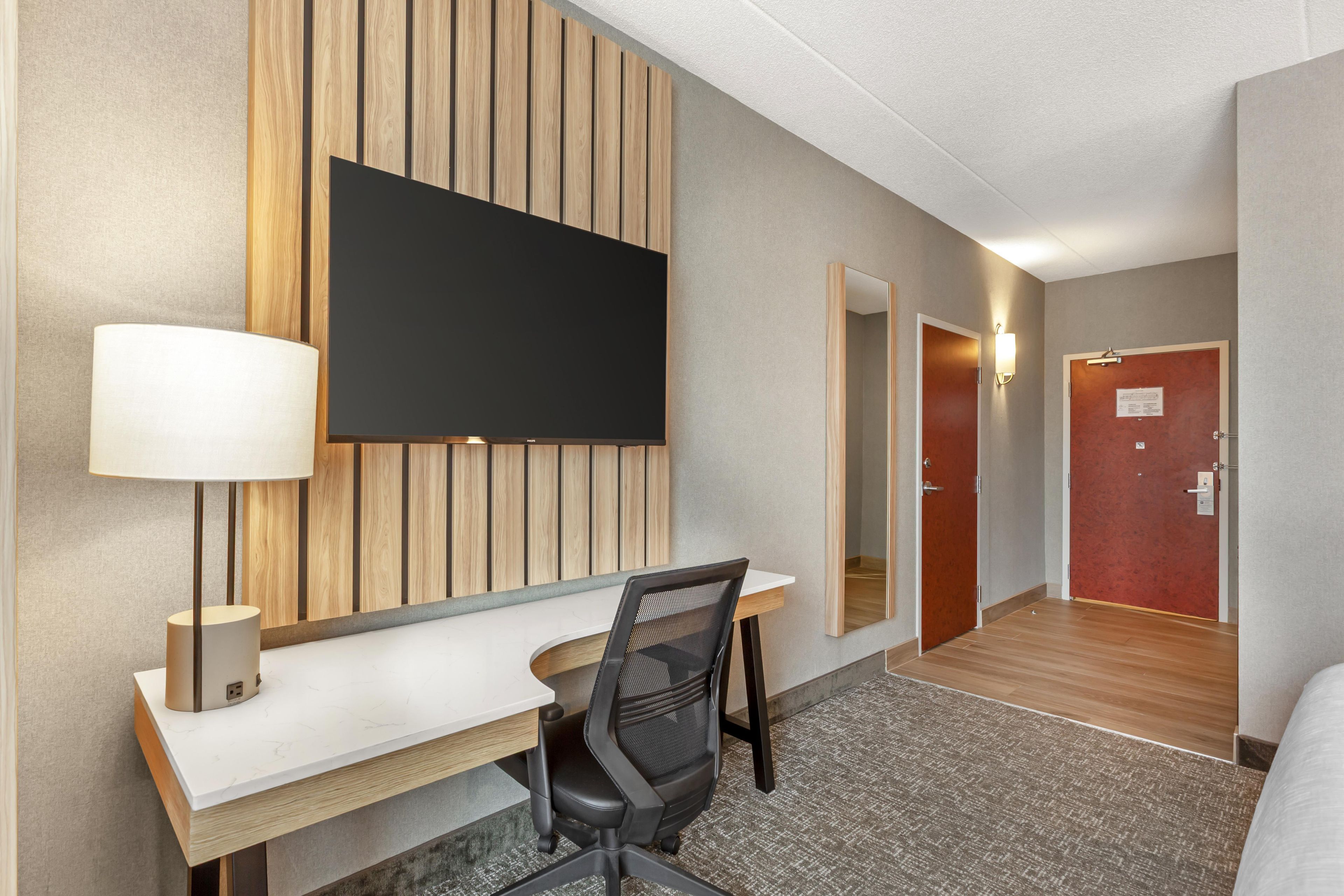 A hotel room with a desk, chair, television, and a mirror.