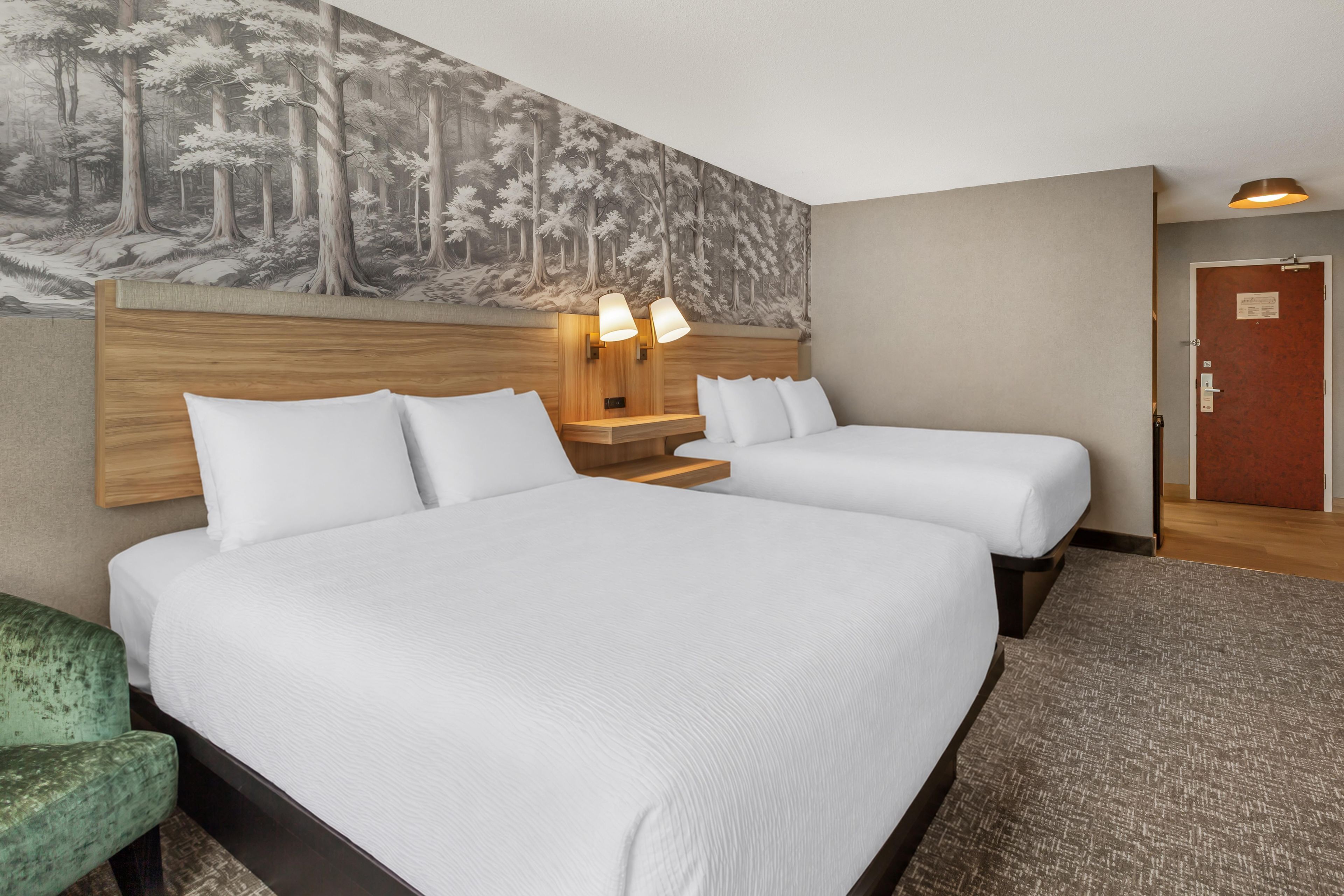Habitación de hotel con dos camas, cabecera con un mural de bosque, lámparas de noche y una puerta con un panel rojo.