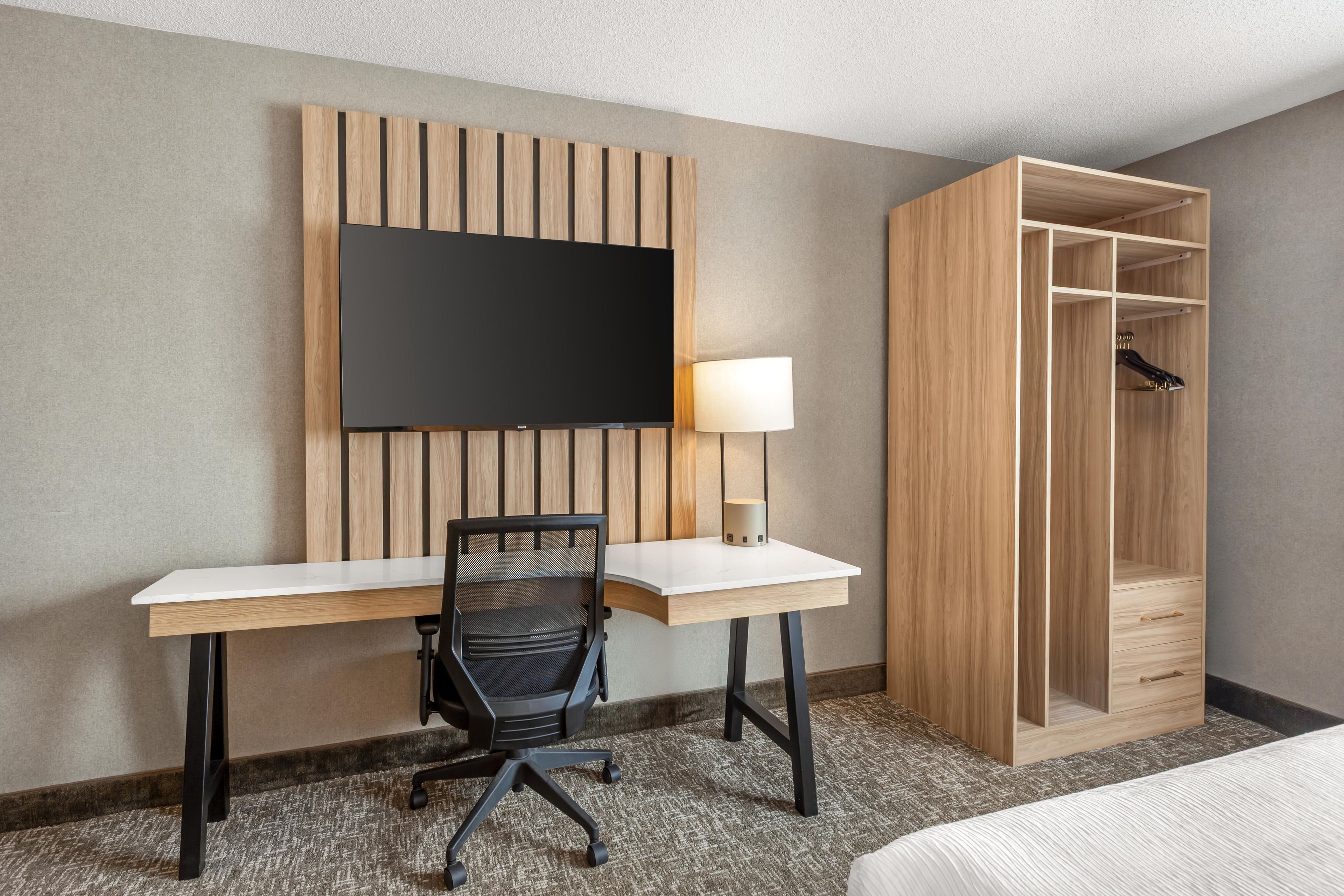 A hotel room with a desk, chair, TV, lamp, and wardrobe.