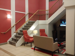 A spacious lobby with a staircase, red and beige chairs, a TV, and a fireplace.