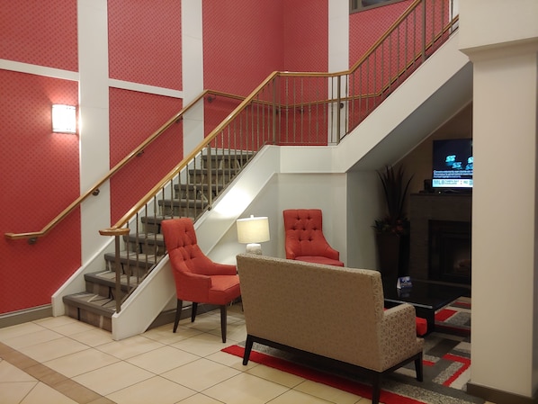 A spacious lobby with a staircase, red and beige chairs, a TV, and a fireplace.