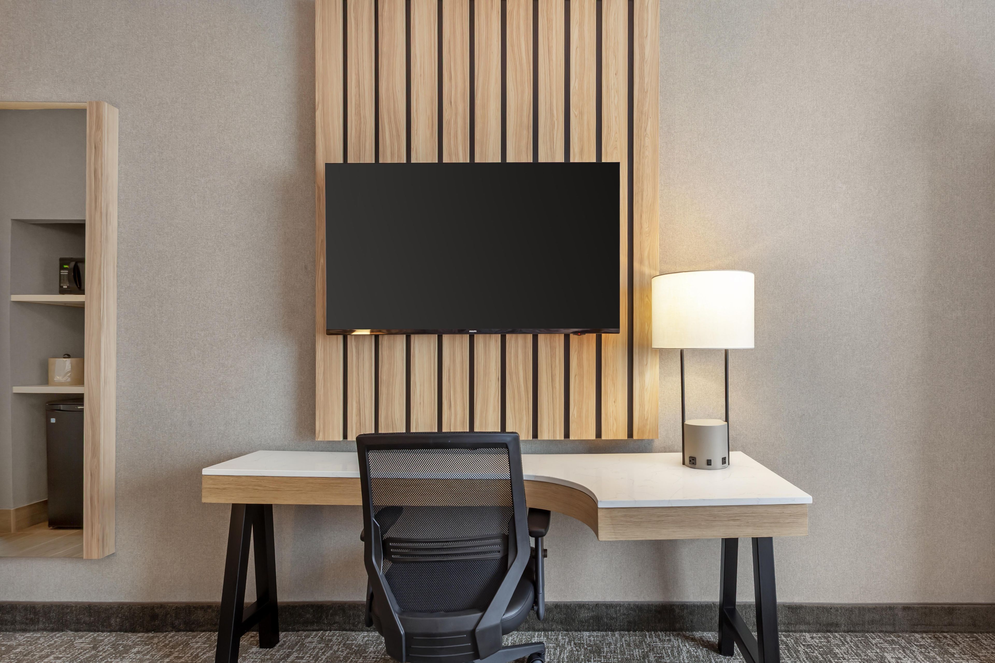 A hotel room with a flat-screen TV mounted on a wooden panel wall, a desk with a lamp, and a black office chair.