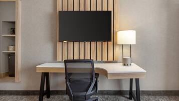 A hotel room with a flat-screen TV mounted on a wooden panel wall, a desk with a lamp, and a black office chair.