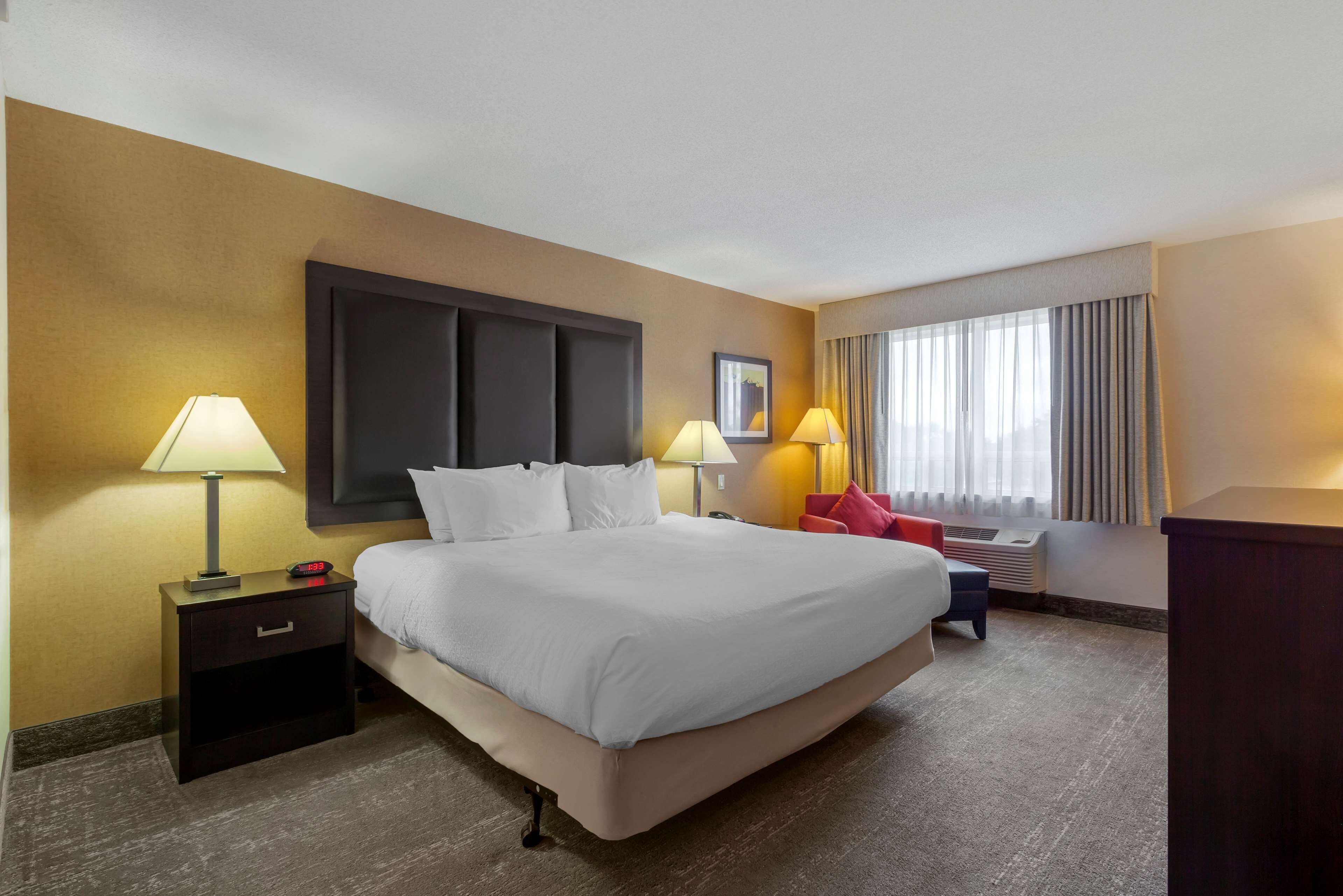 A hotel room with a large bed, two bedside tables with lamps, a red chair, and a window with curtains.