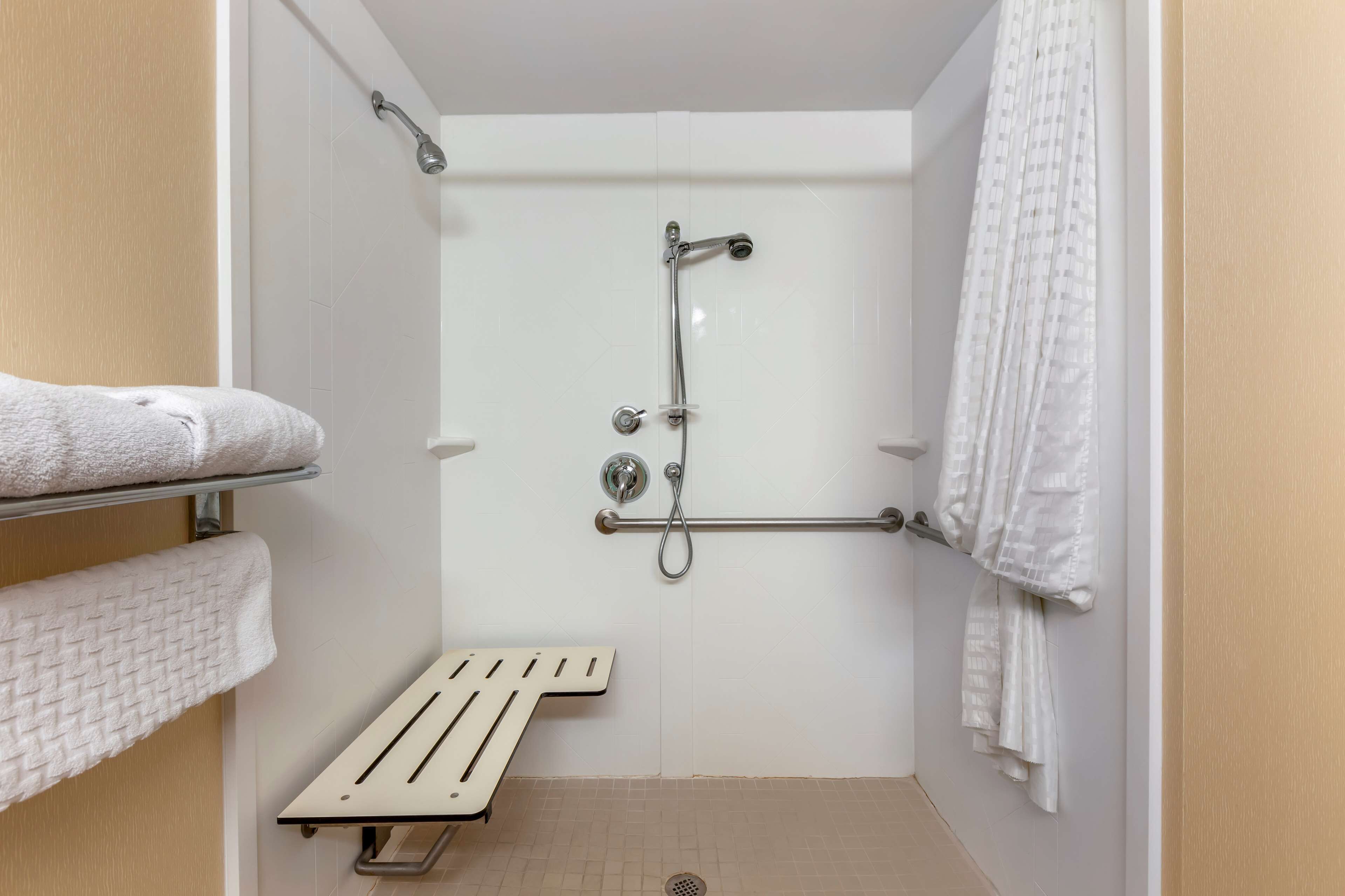 Pequeño espacio de ducha cerrado, de color blanco, con barra de apoyo, toallero y ducha.