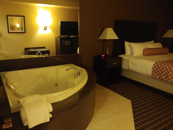 A hotel room with a large bathtub, a bed with white linens, two bedside lamps, and a nightstand with a clock.