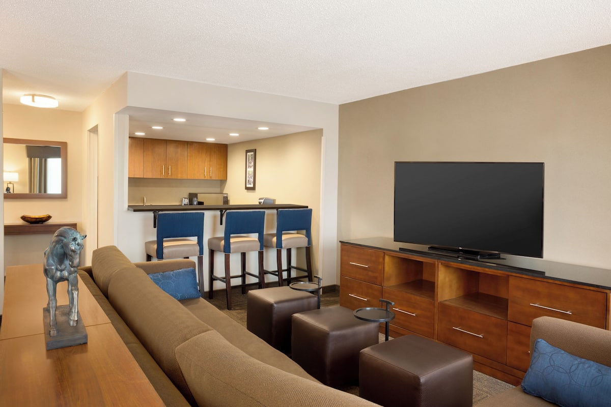 Una sala de estar moderna con sofá, otomanas, televisor y una zona de cocina con taburetes de bar.