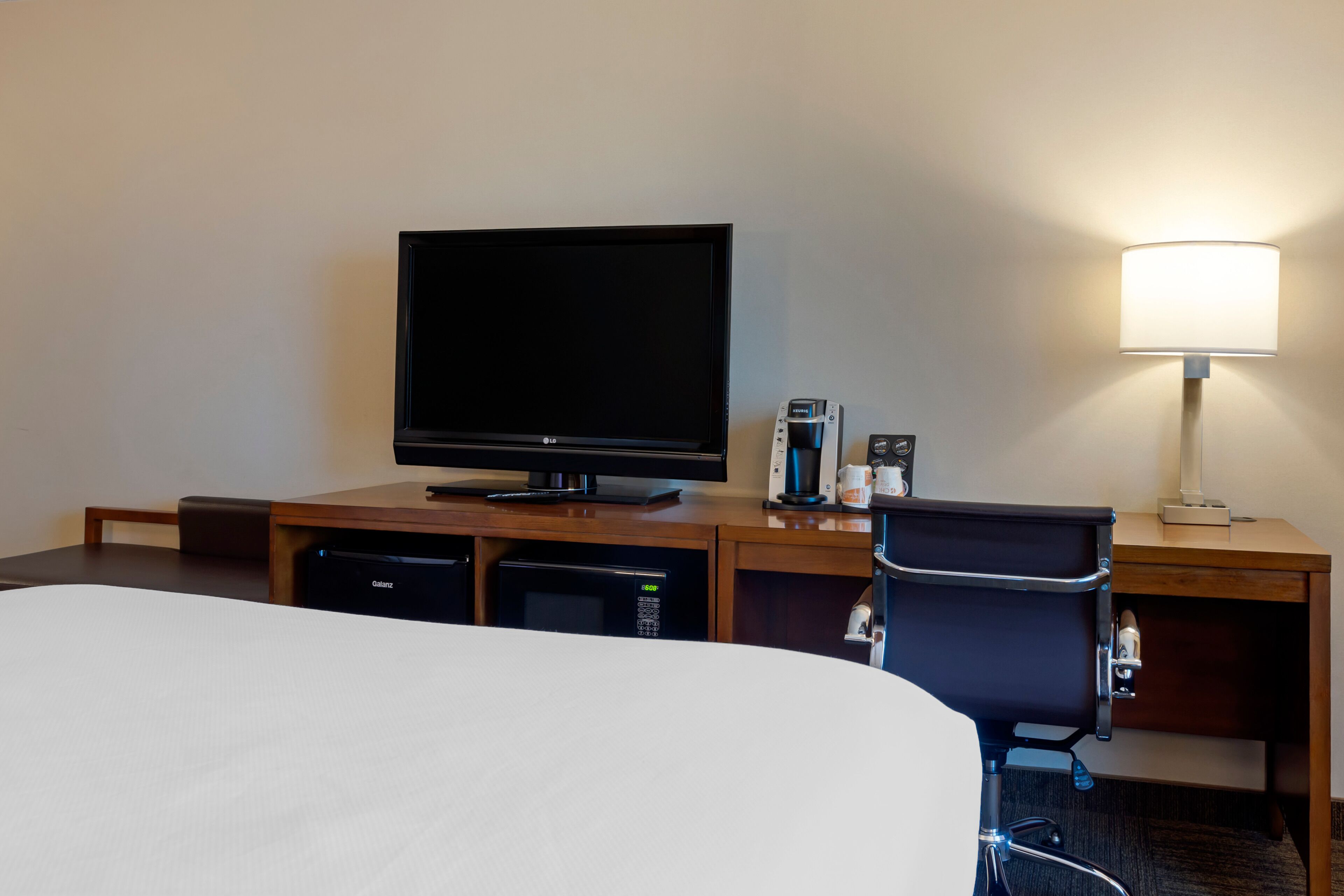Quarto de hotel com cama, televisão, escrivaninha com luminária, cafeteira e computador.