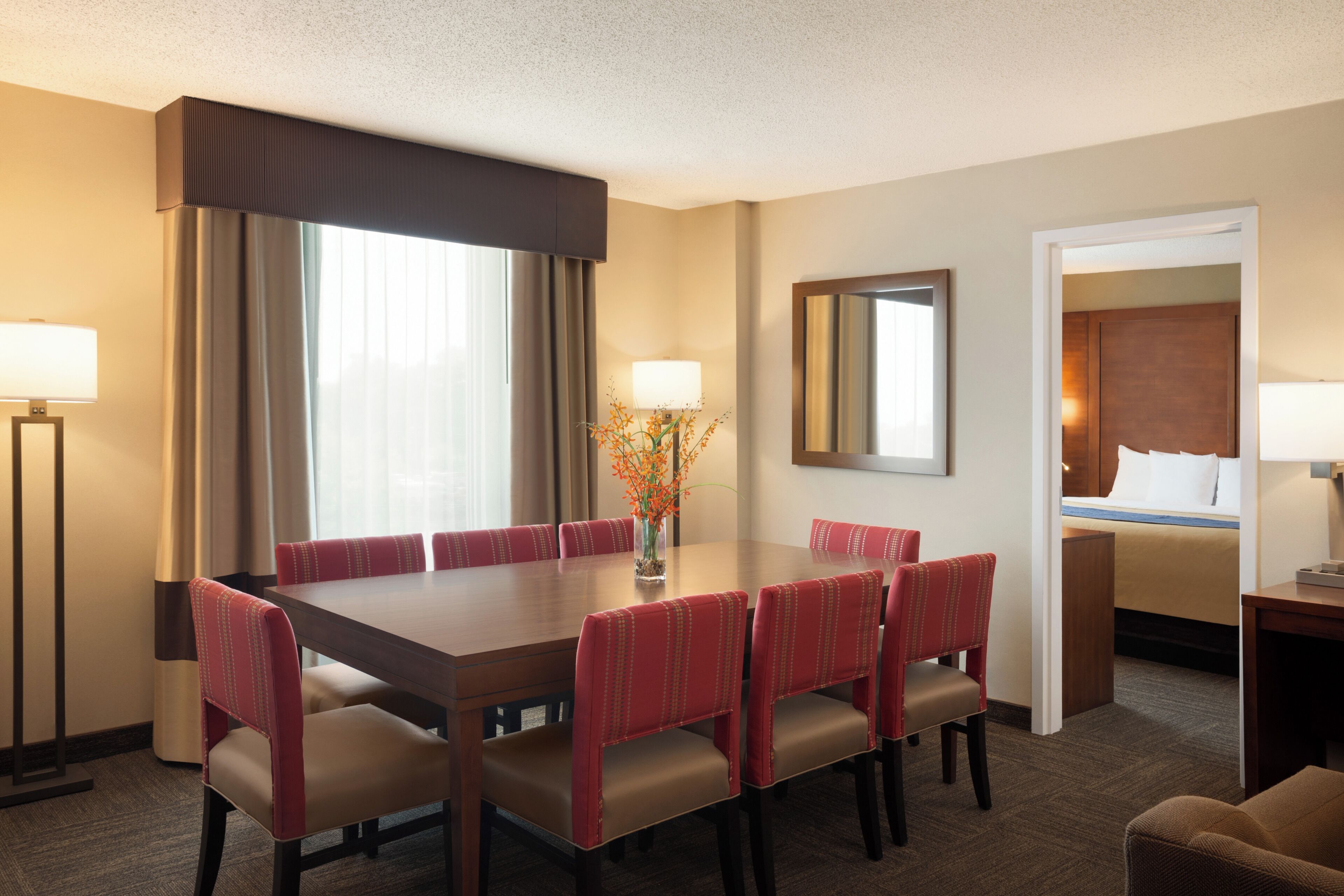 A hotel room with a large dining table, red upholstered chairs, a bed with white linens, a mirror on the wall, and a vase with flowers on the table.