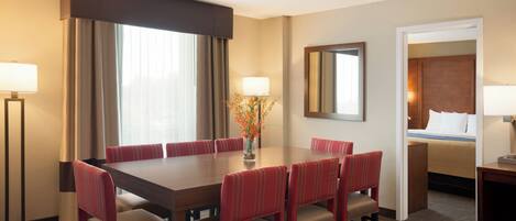 A hotel room with a large dining table, red upholstered chairs, a bed with white linens, a mirror on the wall, and a vase with flowers on the table.