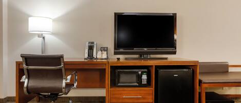 A hotel room with a desk, chair, flat-screen TV, microwave, mini-fridge, and a lamp.