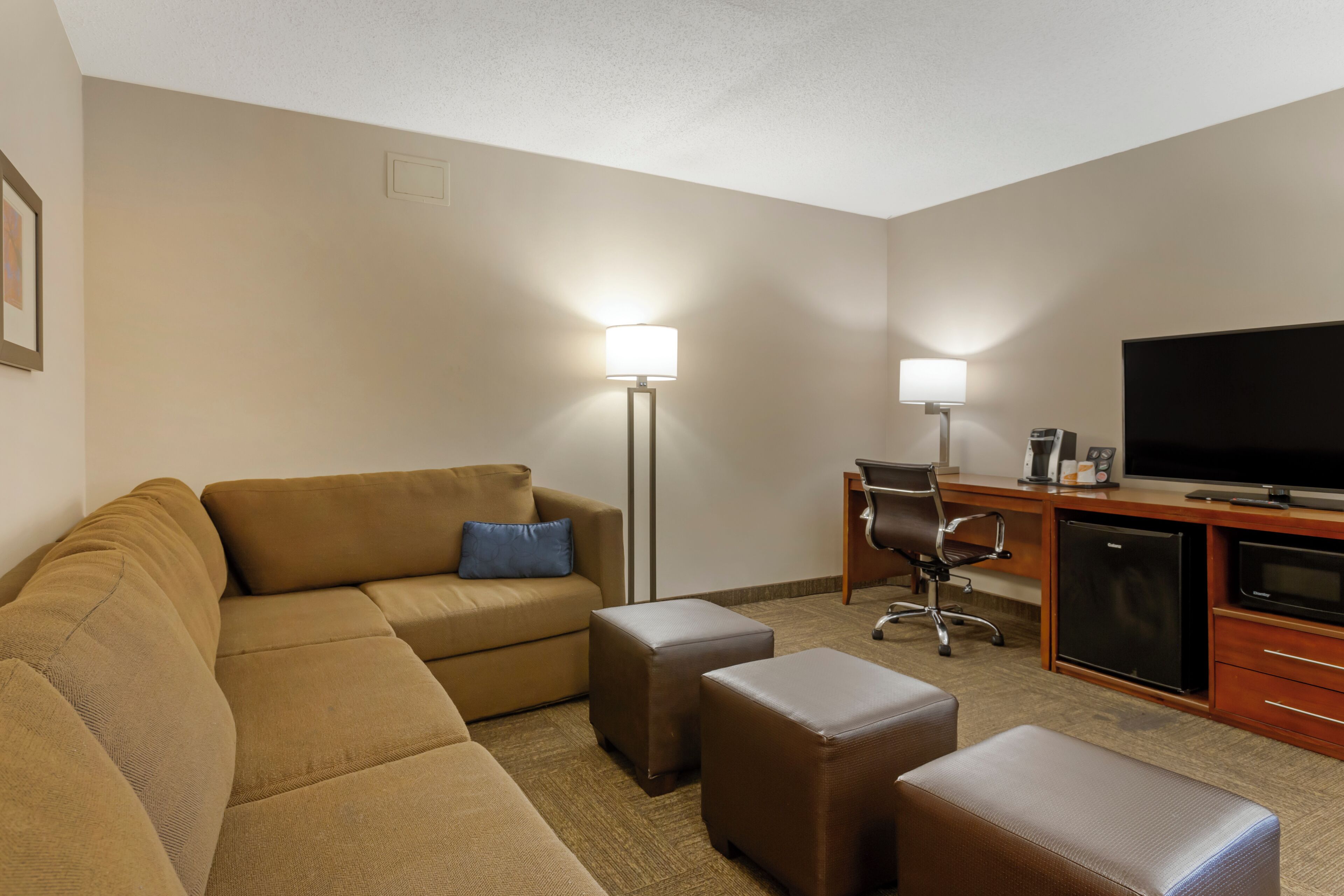 A hotel room with a sofa, ottomans, a desk with a television, and two floor lamps.