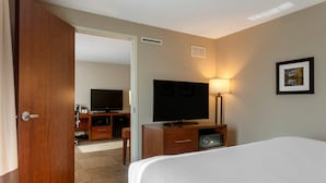 A hotel room with a bed, a TV, a desk with a lamp, and a door leading to another room.