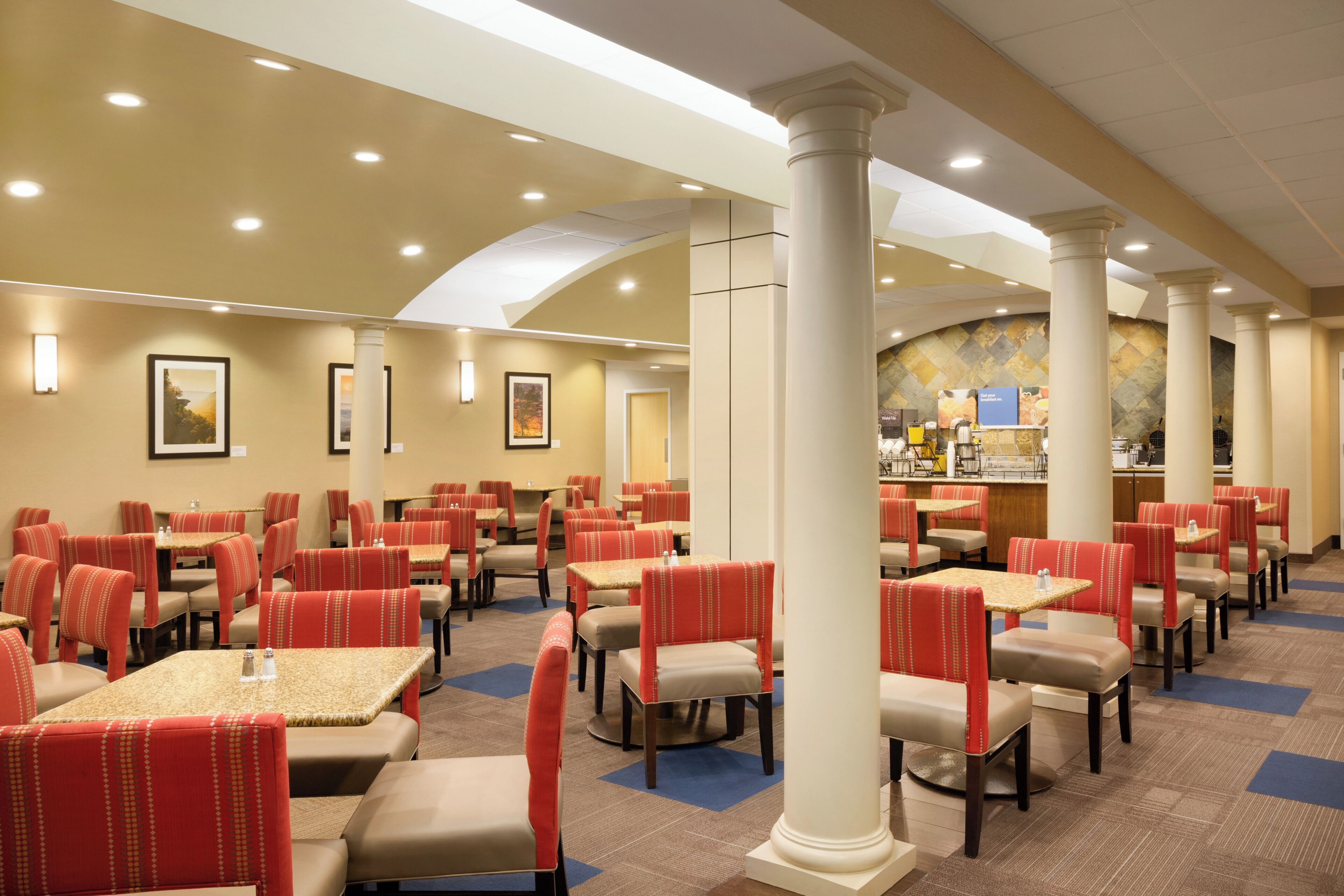 A hotel dining area with multiple tables and chairs, a central column, and a counter with food items.