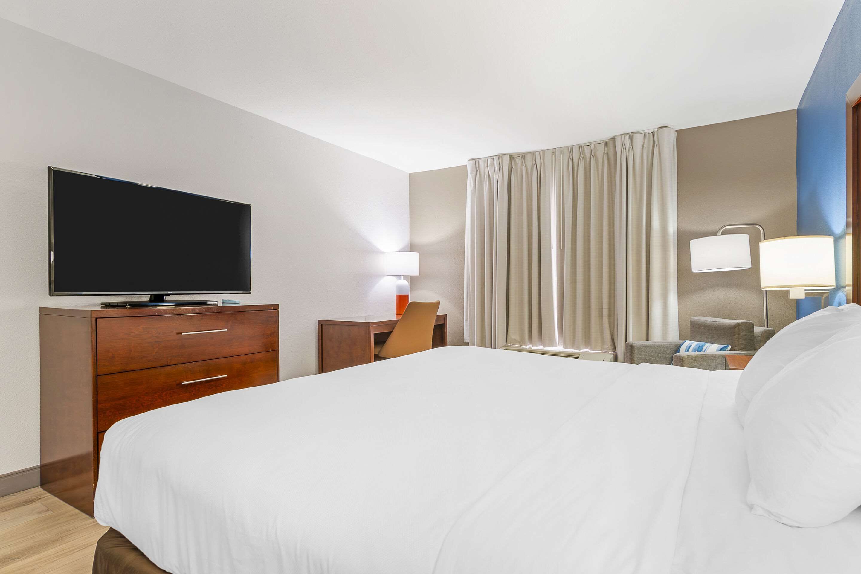 Una habitación de hotel con una cama grande, un televisor sobre un aparador de madera, un escritorio con una silla, una lámpara y una ventana con cortinas.