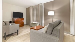 A modern hotel room with a grey sofa, a wooden coffee table, a flat-screen TV, a wooden dresser, and a large window with curtains. - Comfort Inn Near Six Flags St. Louis (Pacific)