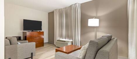 A modern hotel room with a grey sofa, a wooden coffee table, a flat-screen TV, a wooden dresser, and a large window with curtains.
