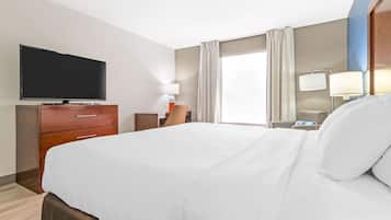 A hotel room with a large bed, a flat-screen TV, a wooden dresser, a chair, a desk with a lamp, and a window with curtains.