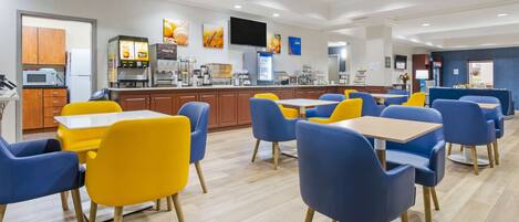 A modern dining area with yellow and blue chairs, a flat-screen TV, and a kitchen area with appliances.