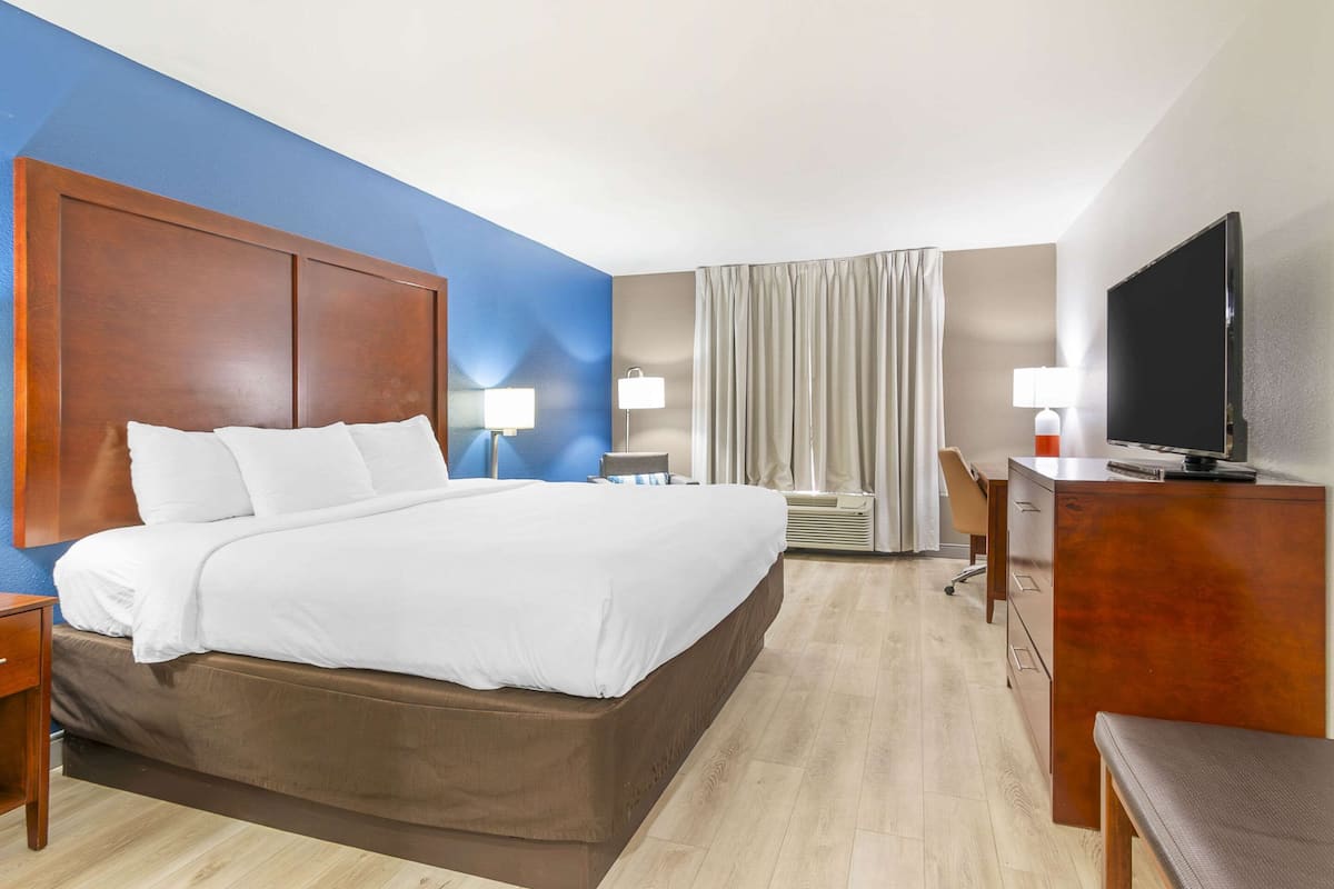 A hotel room with a large bed, bedside tables, a desk, a chair, a television, and a window with curtains.
