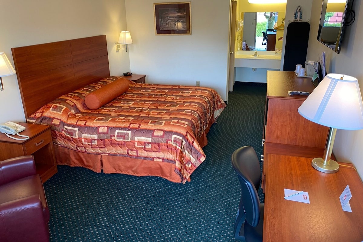 Une chambre d’hôtel avec un grand lit, un bureau, une chaise, une lampe de bureau, un téléphone, un miroir et un ventilateur de plafond.