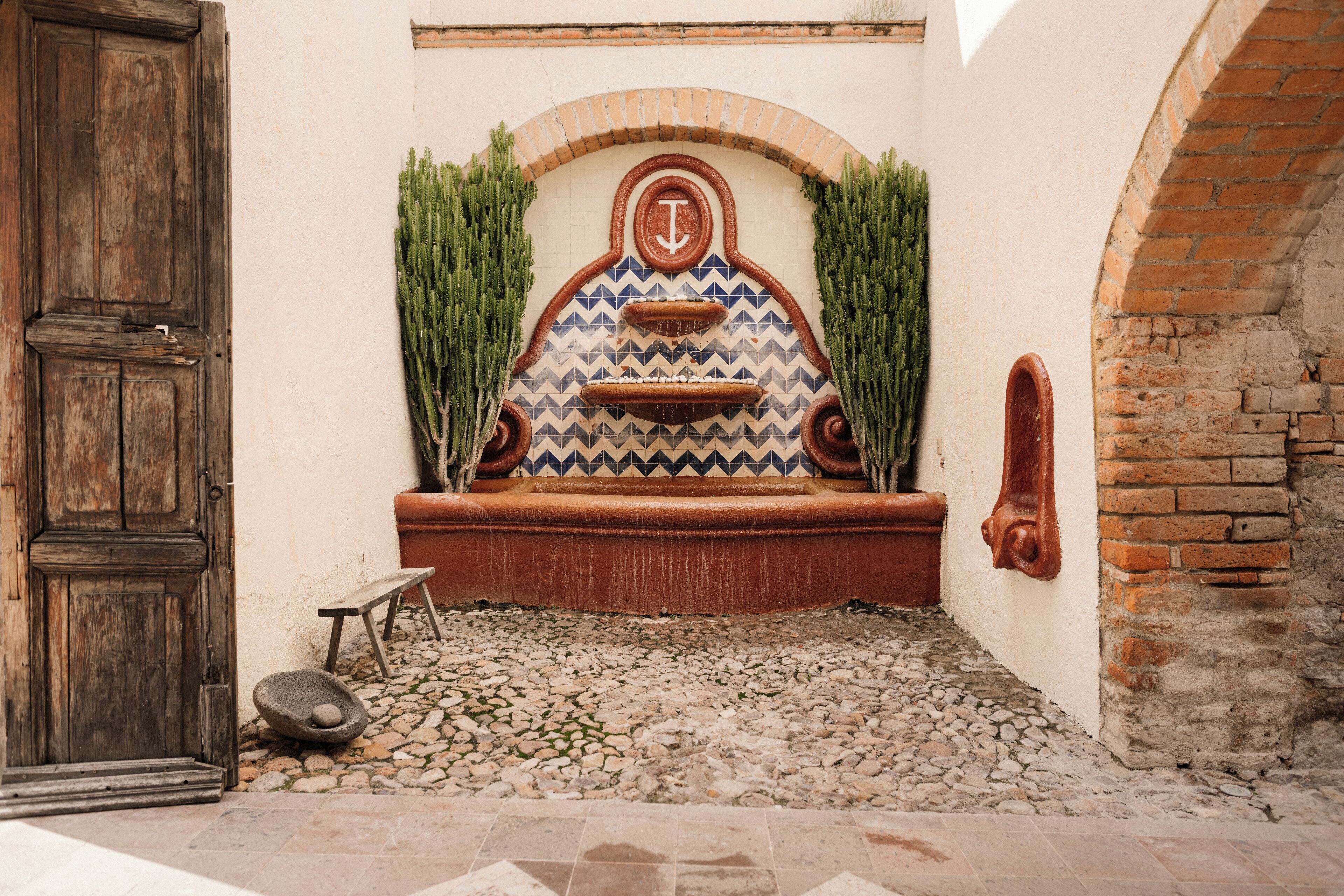 A courtyard with a rustic wooden door, a stone bench, and a tiled wall with a geometric pattern and a central anchor symbol.