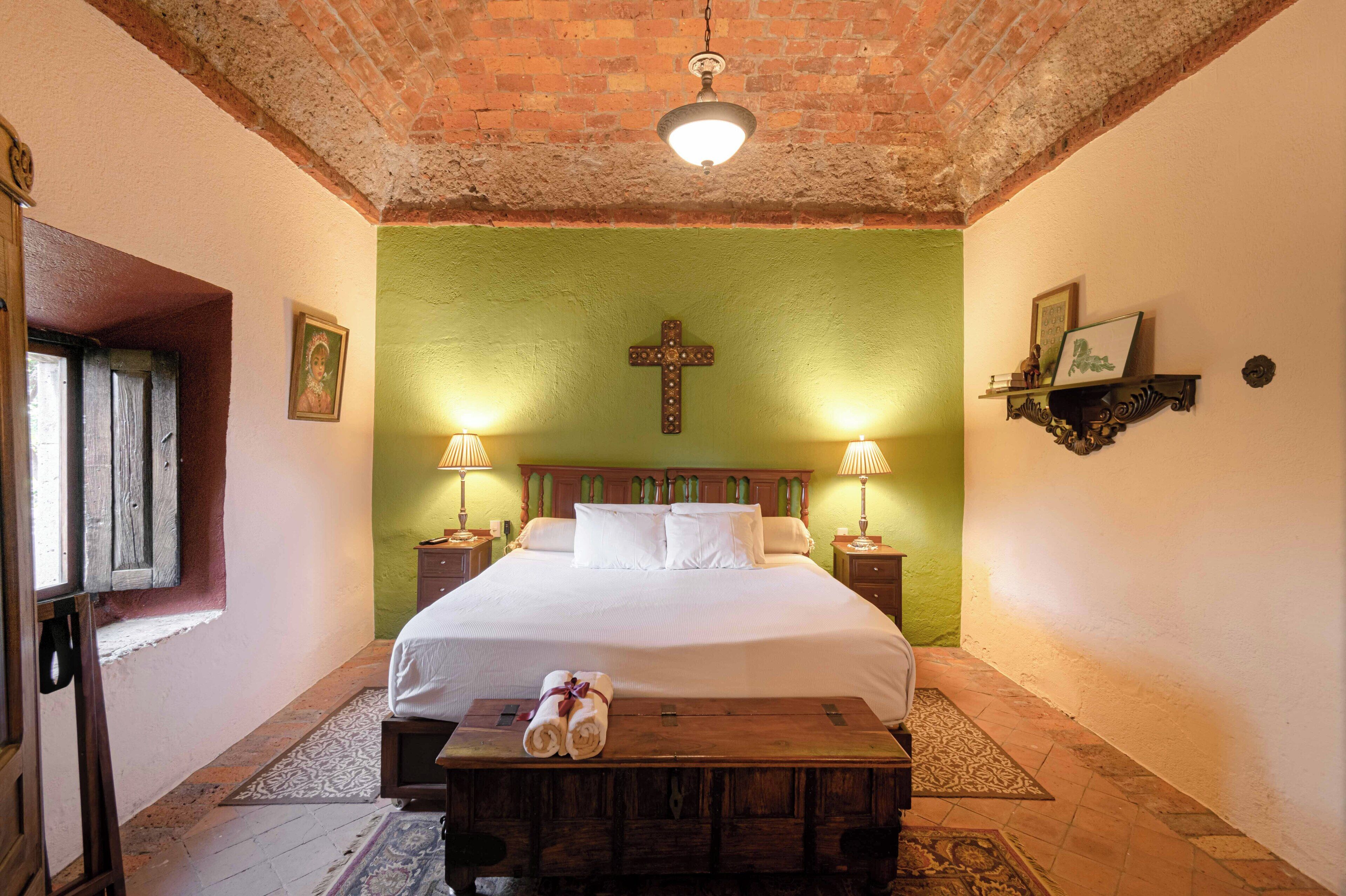 A room with a large bed, two bedside lamps, a cross on the wall, a wooden bench with towels, and a brick ceiling.