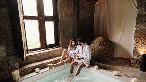 Un couple est assis près d’une baignoire en pierre, entouré de bougies, dans une pièce rustique dotée d’une grande fenêtre et d’un panier en osier.