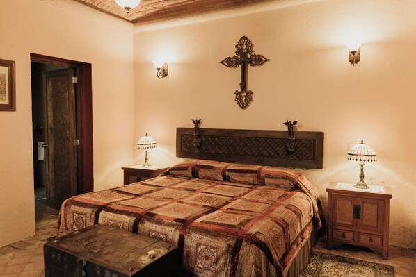 A traditional bedroom with a large bed, wooden headboard, two bedside tables with lamps, a wooden bench, and a cross on the wall.