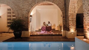 Un espace piscine agrémenté d’arches en pierre, un couple assis à une table éclairée par des bougies et une plante en pot.