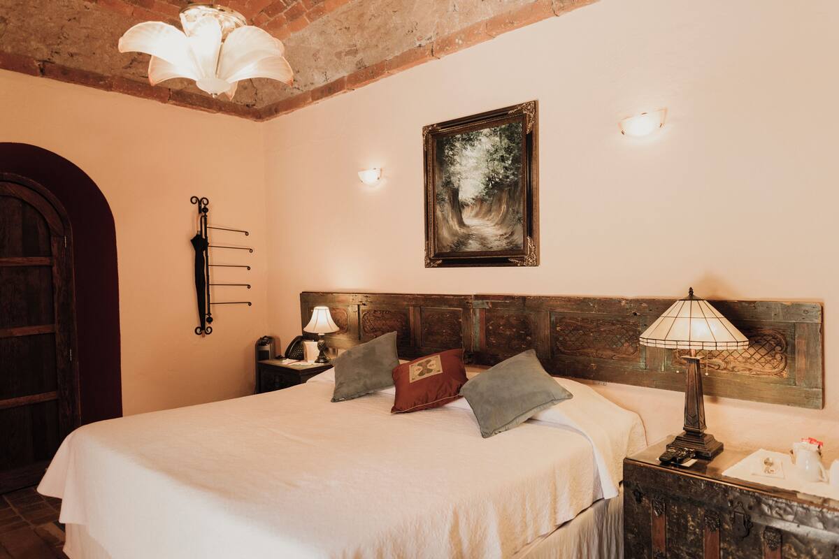 Ein Schlafzimmer mit Bett, Nachttisch, Lampe, einem an der Wand hängenden Bild und einer Holztür.