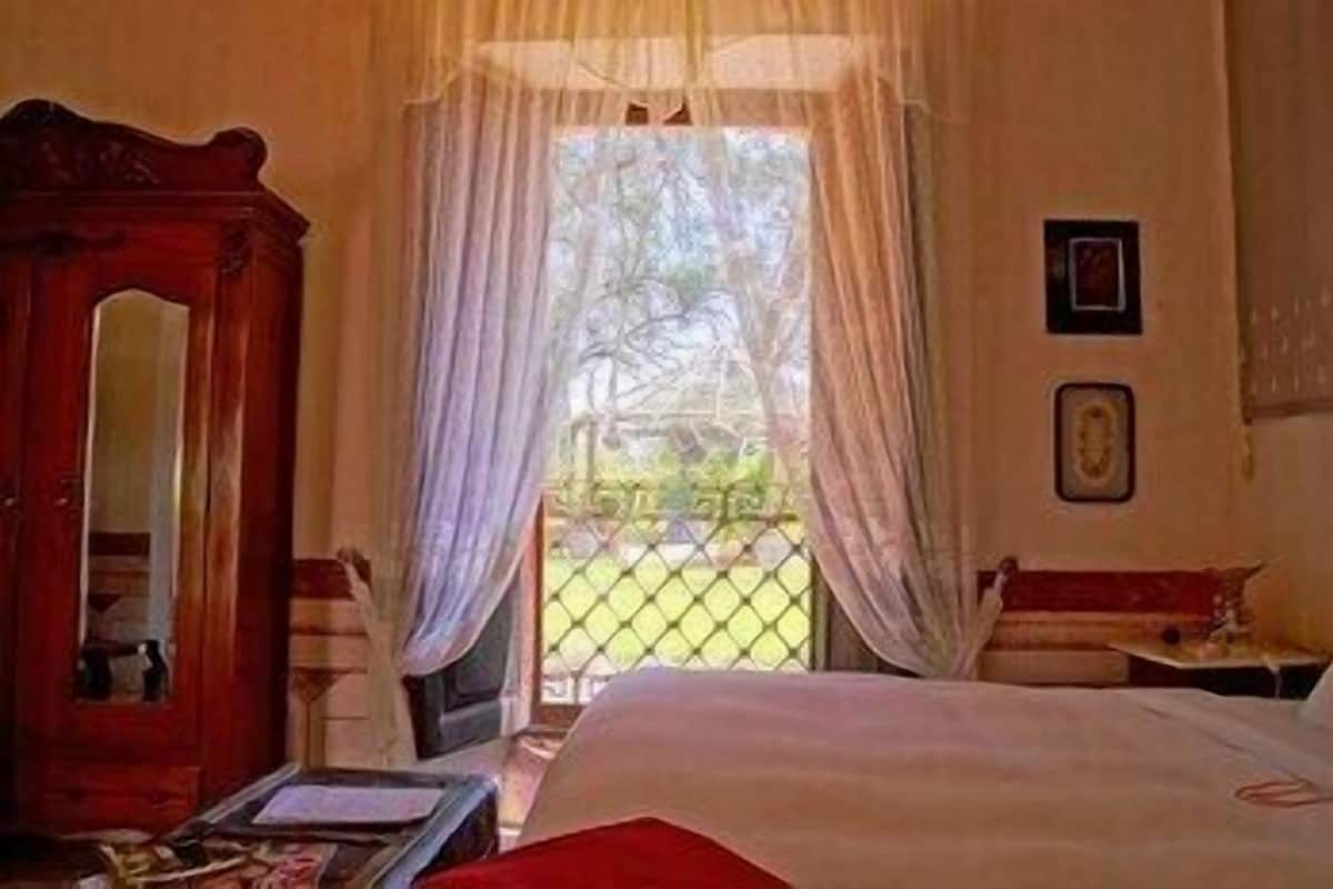 Ein Zimmer mit einem Bett, einem Holzschrank, einem Fenster mit durchsichtigen Vorhängen und einem kleinen Tisch mit einer Lampe.