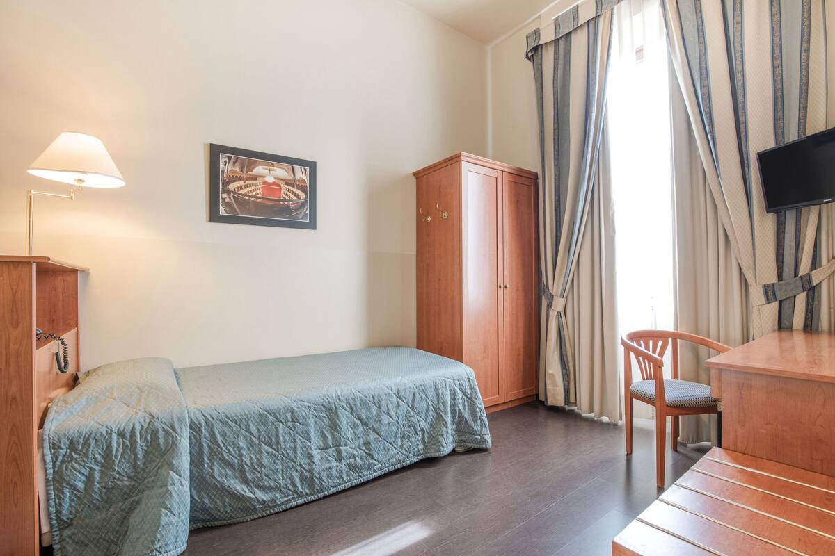 Una habitación con cama, escritorio, televisor, armario y ventana con cortinas.