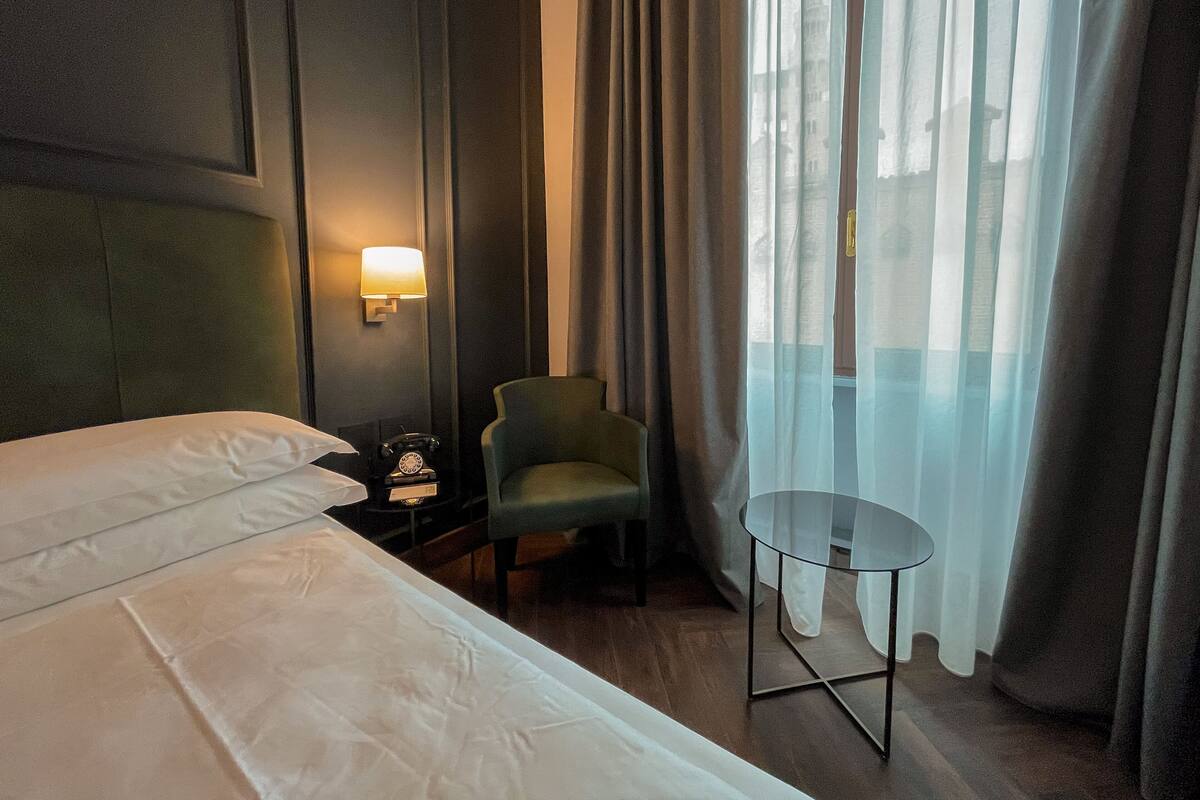 Habitación de hotel con cama, una silla, una mesita, una lámpara y un ventanal con cortinas.