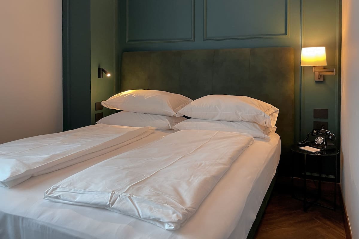 Una cama bien hecha con sábanas y almohadas blancas, una lámpara de noche y un teléfono sobre una mesita junto a la cama.