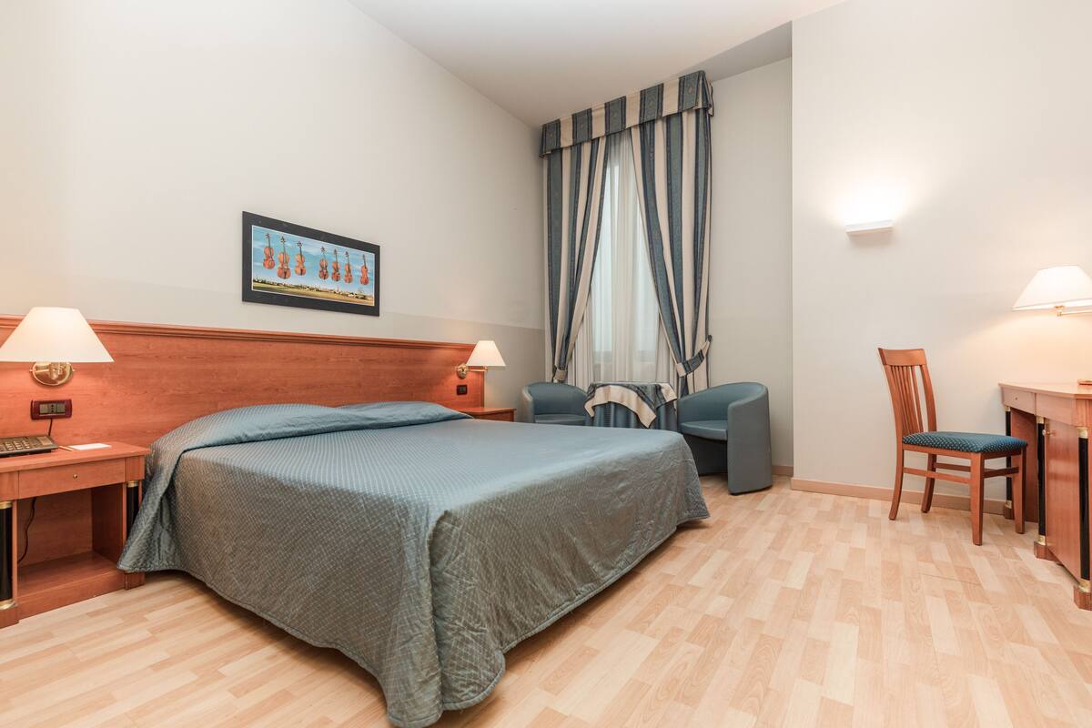 Una habitación de hotel con una cama, dos mesitas de noche, un escritorio, una silla, una lámpara y una cortina.