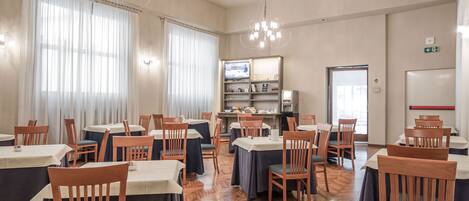 A hotel dining area with tables and chairs, a television, and a kitchen in the background.