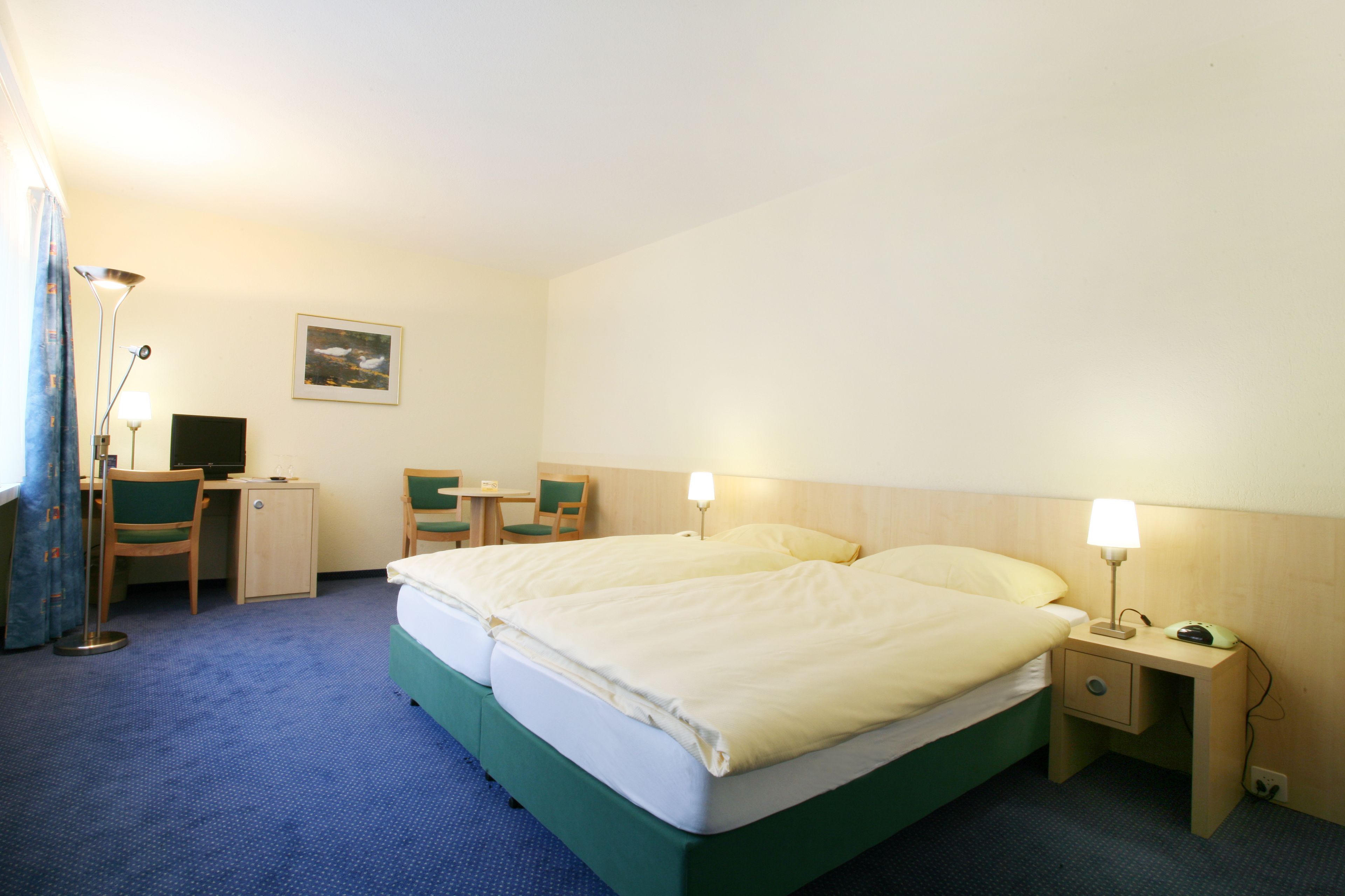 A hotel room with a double bed, a desk with a chair, a small table, and a nightstand with a lamp.