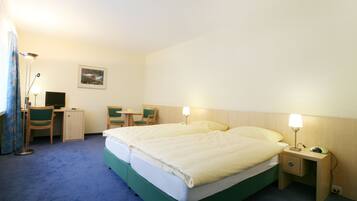 A hotel room with a double bed, a desk with a chair, a small table, and a nightstand with a lamp.