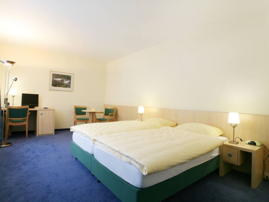 A hotel room with a double bed, a desk with a chair, a small table, and a nightstand with a lamp.