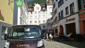 Ein Catering-Van parkt auf einer Kopfsteinpflasterstraße, im Hintergrund sind Gebäude zu sehen, darunter ein Hotel und ein Restaurant.