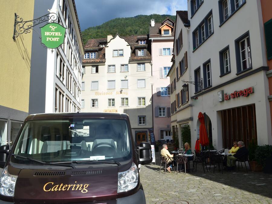 A catering van parked on a cobblestone street with buildings in the background, including a hotel and a restaurant.
