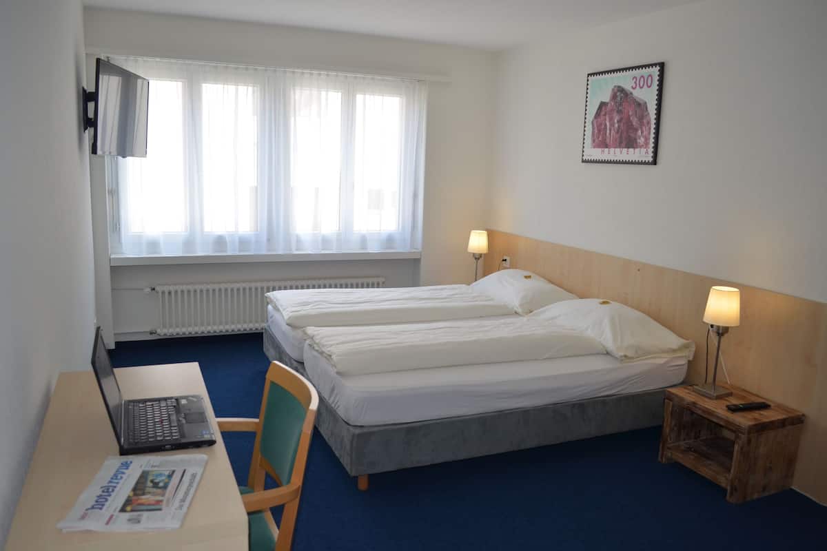 A hotel room with two beds, a desk with a laptop, a chair, a nightstand with a lamp, and a window with sheer curtains.
