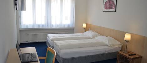 A hotel room with two beds, a desk with a laptop, a chair, a nightstand with a lamp, and a window with sheer curtains.