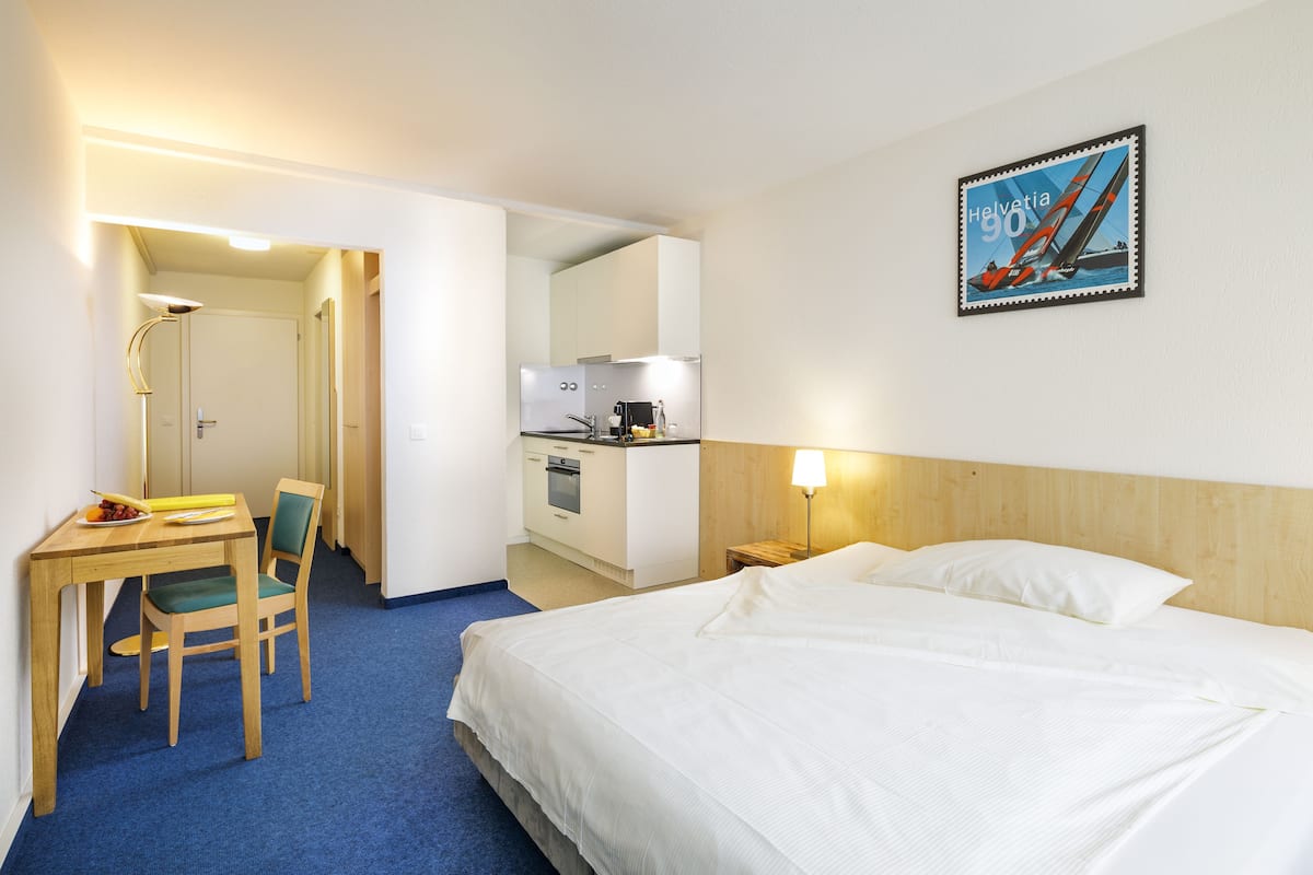A hotel room with a bed, a desk with a chair, a small kitchenette, and a mirror.