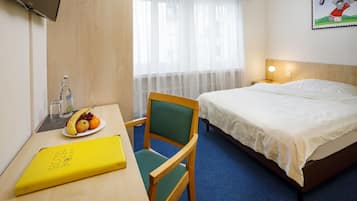 Quarto de hotel com cama, escrivaninha com cadeira, garrafa de água, prato com fruta e quadro na parede.